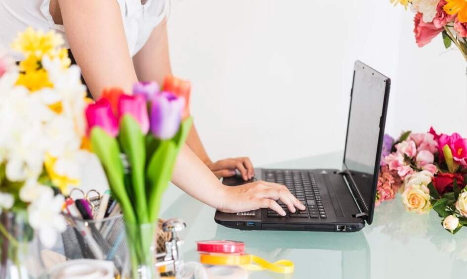 A female florists checking out for flower orders online on a laptop, with flowers sitting around.